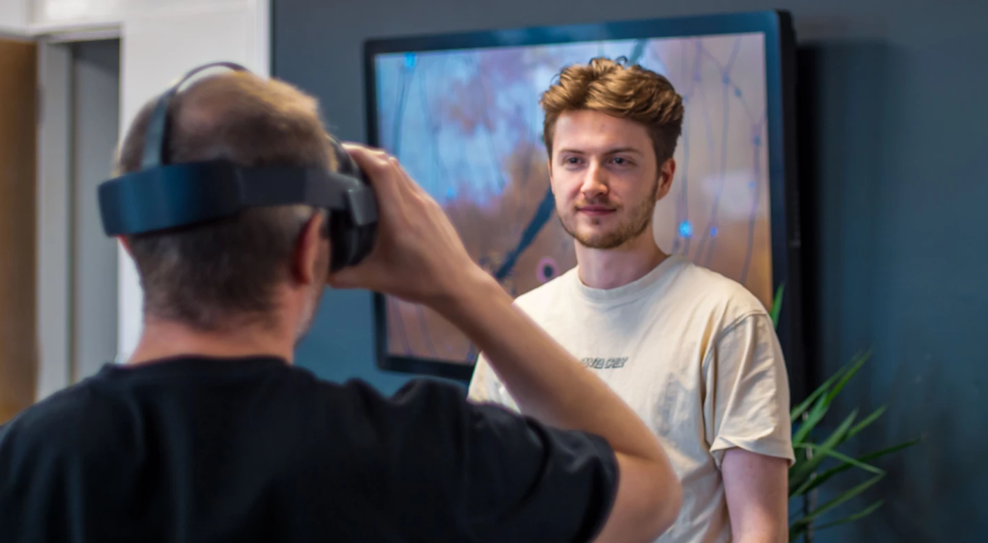 A man in a white t-shirt observing someone using a virtual reality headset