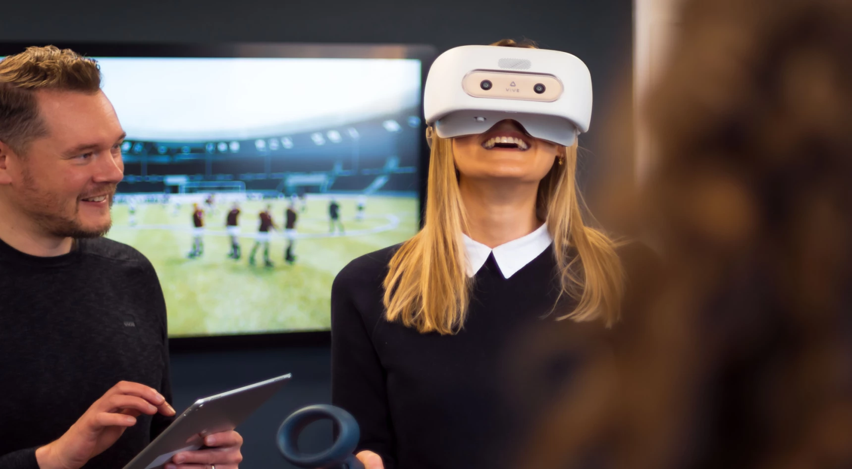 a smiling woman in a VR headset being supervised by a man holding a tablet
